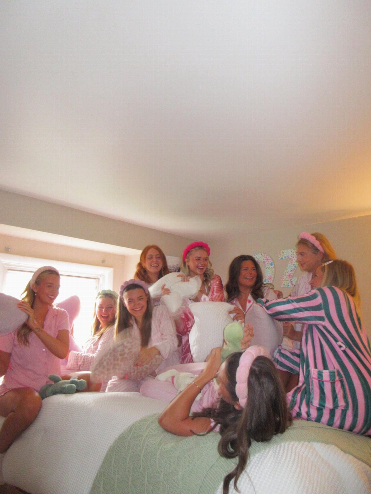 Teenage girls in pink pajamas having a pillow fight on a white bed.