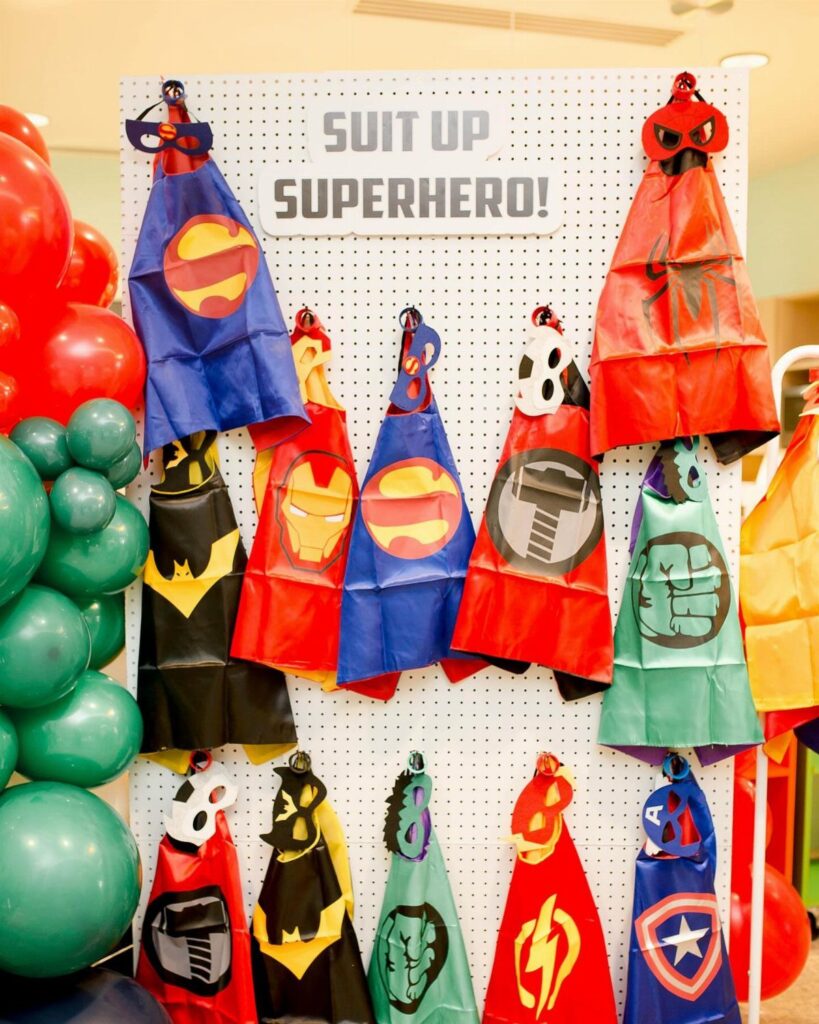 Superhero capes and masks hanging on a white pegboard with colorful balloons.