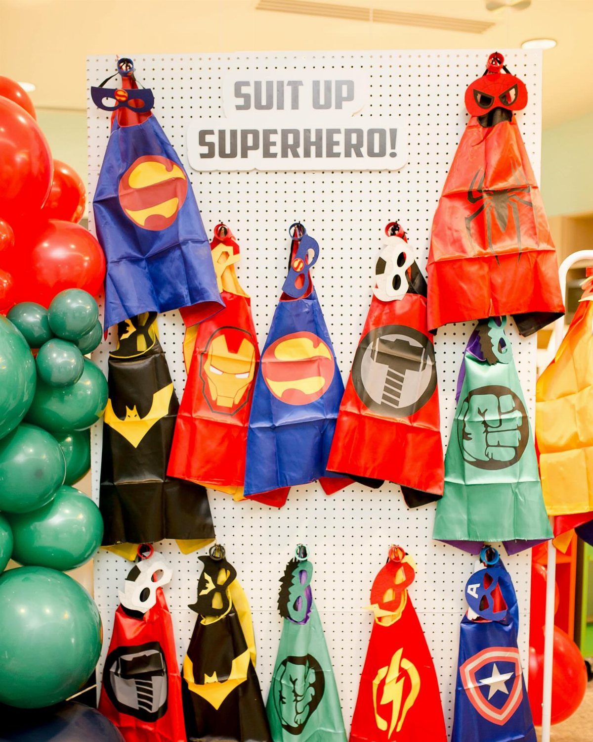 Superhero capes and masks hanging on a white pegboard with colorful balloons.