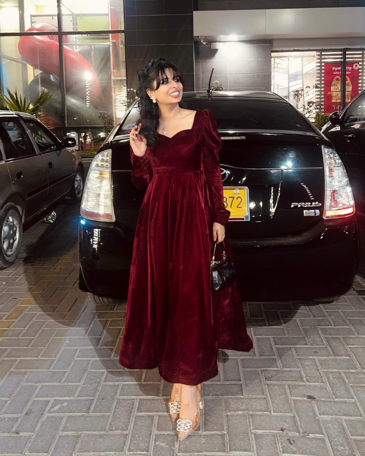 Woman in a long-sleeved burgundy velvet dress posing in front of a black car.