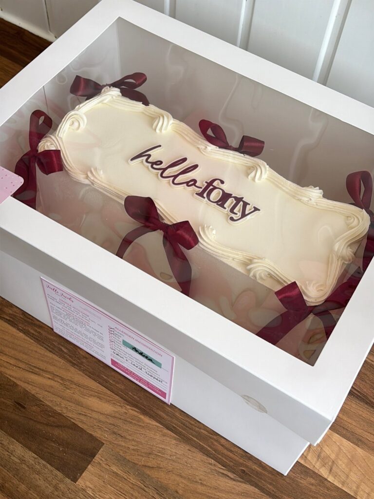 White frosted sheet cake with hello forty text and maroon ribbons in a box