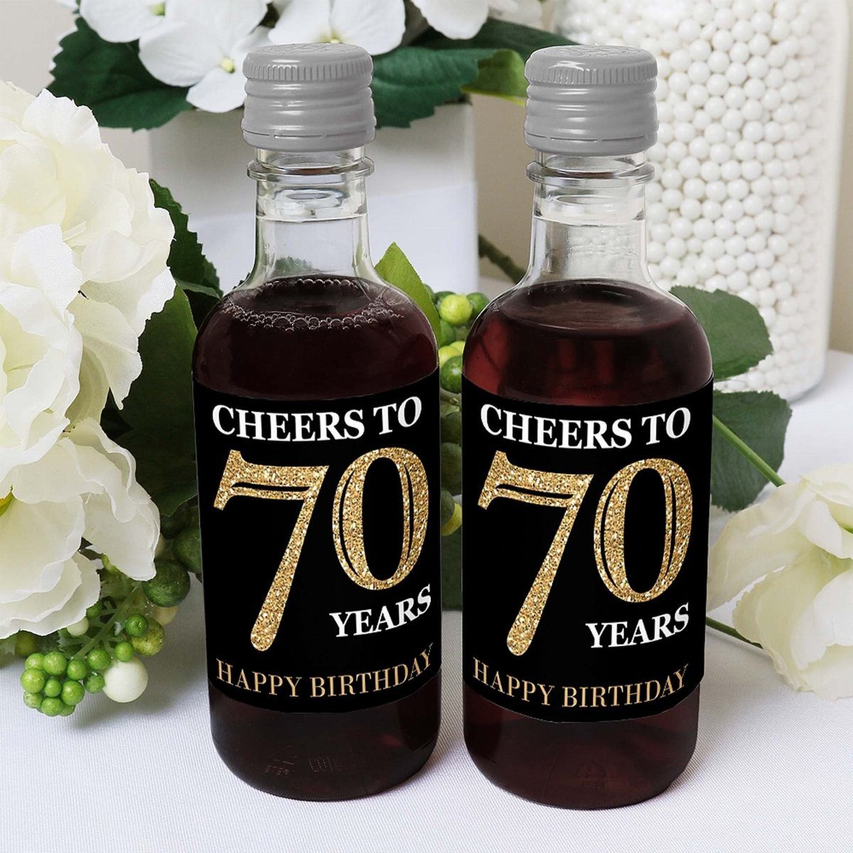 Two mini wine bottles with black and gold 70th birthday labels among white flowers.