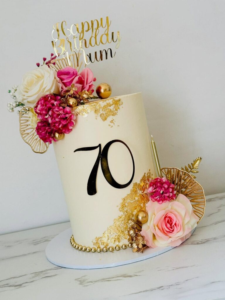 White birthday cake with number 70, pink roses, and gold birthday topper.