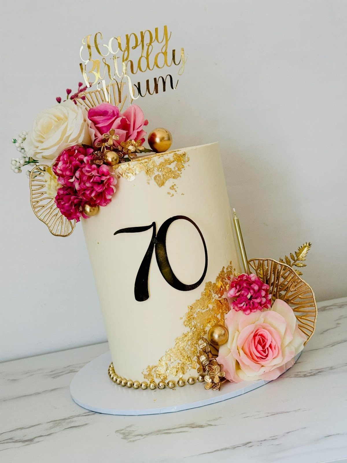 White birthday cake with number 70, pink roses, and gold birthday topper.