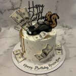 White birthday cake decorated with edible money, a black car, and gold accents.