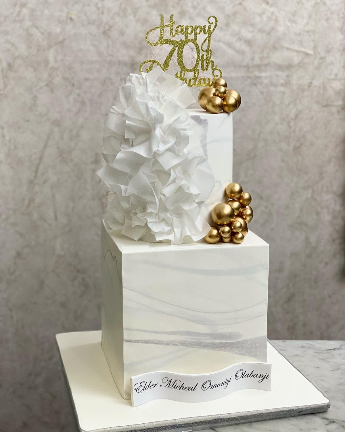 Two-tier white marble cake with gold spheres and a Happy 70th Birthday topper.
