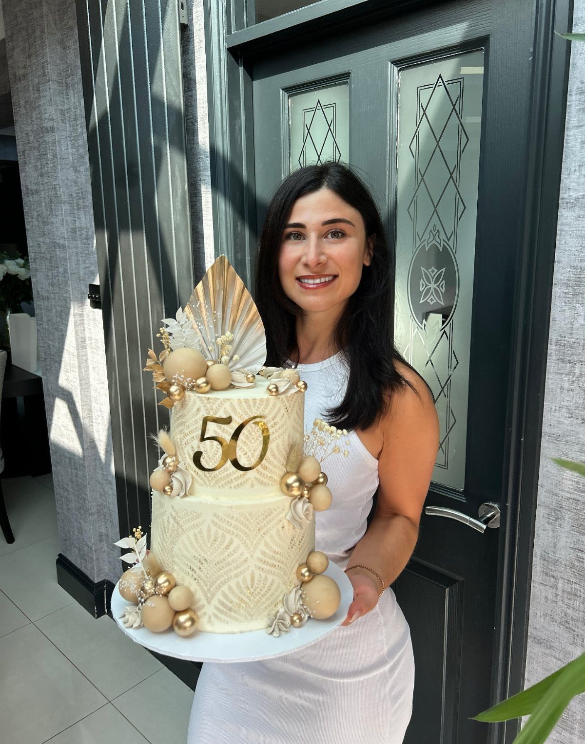 Woman holding a two-tier white and gold 50th birthday cake outdoors