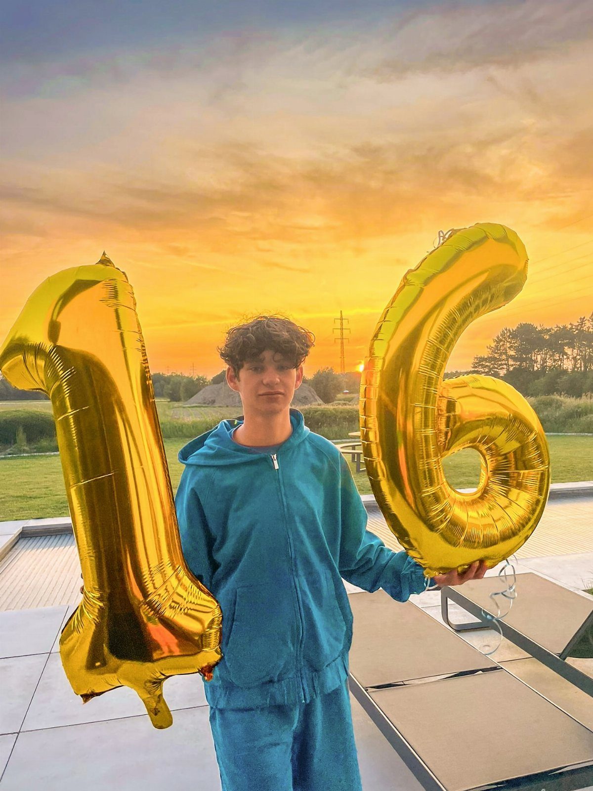 Teenage boy in blue tracksuit holding large gold number 16 balloons at sunset