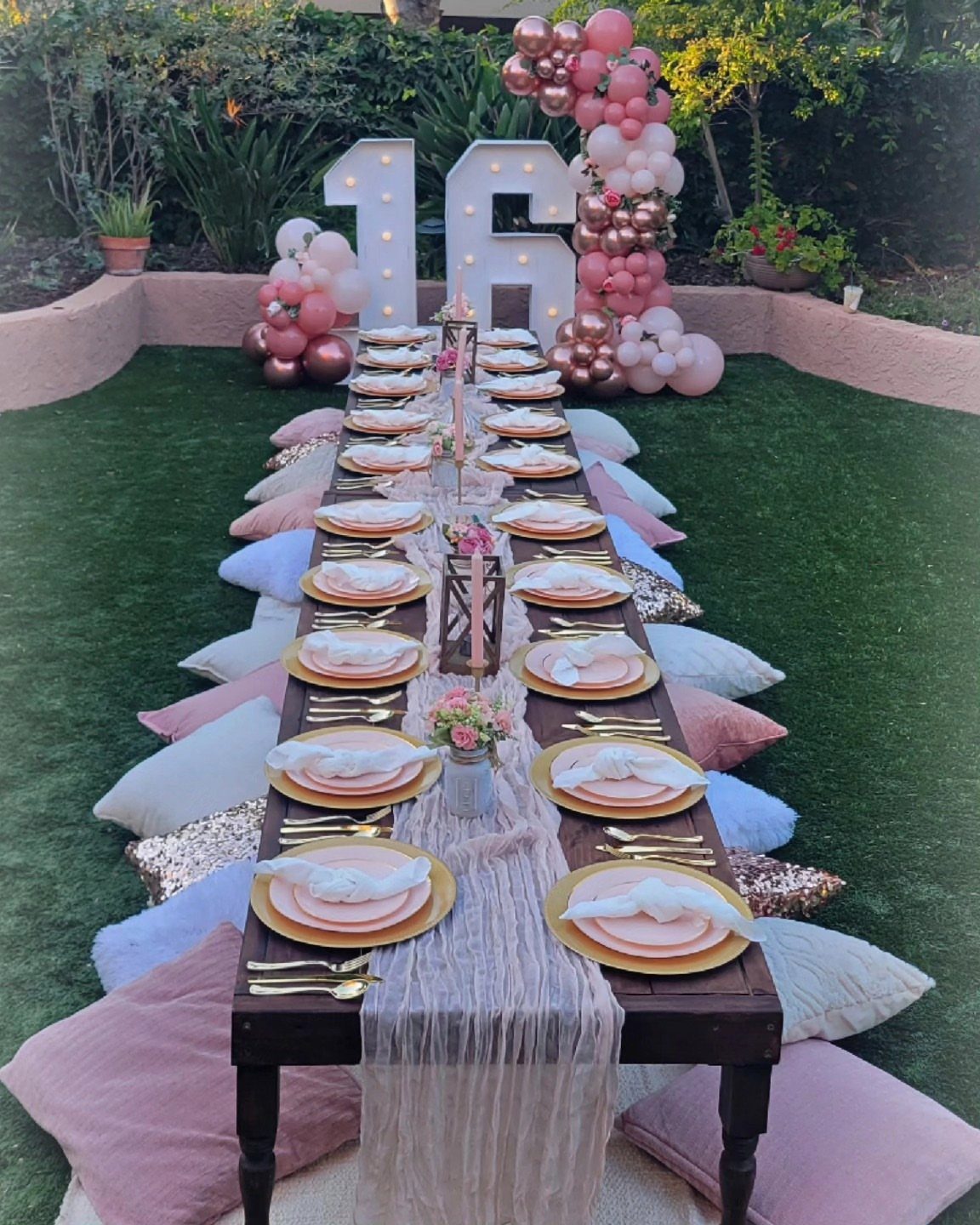 Outdoor party table with gold plates, pink cushions, and large glowing 16 numbers.
