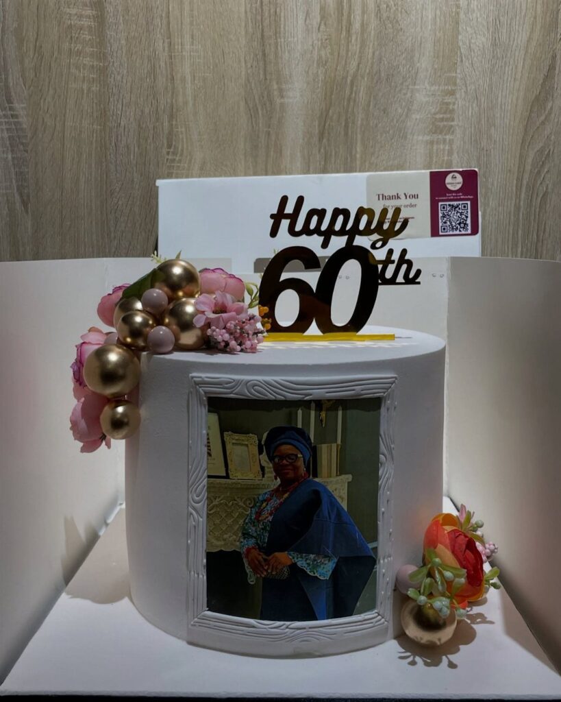 White 60th birthday cake with edible photo, gold topper, and pink floral decorations.