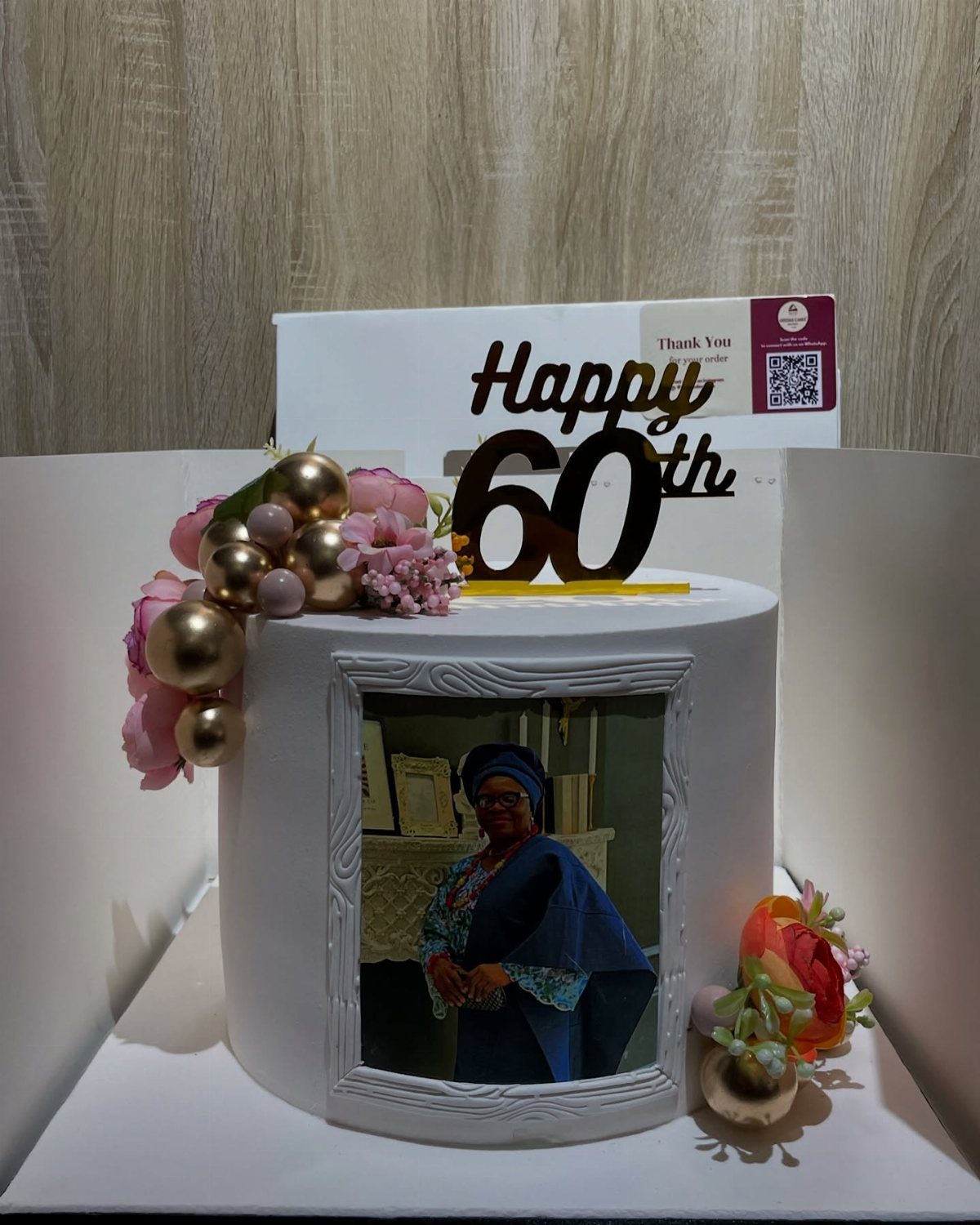 White 60th birthday cake with edible photo, gold topper, and pink floral decorations.