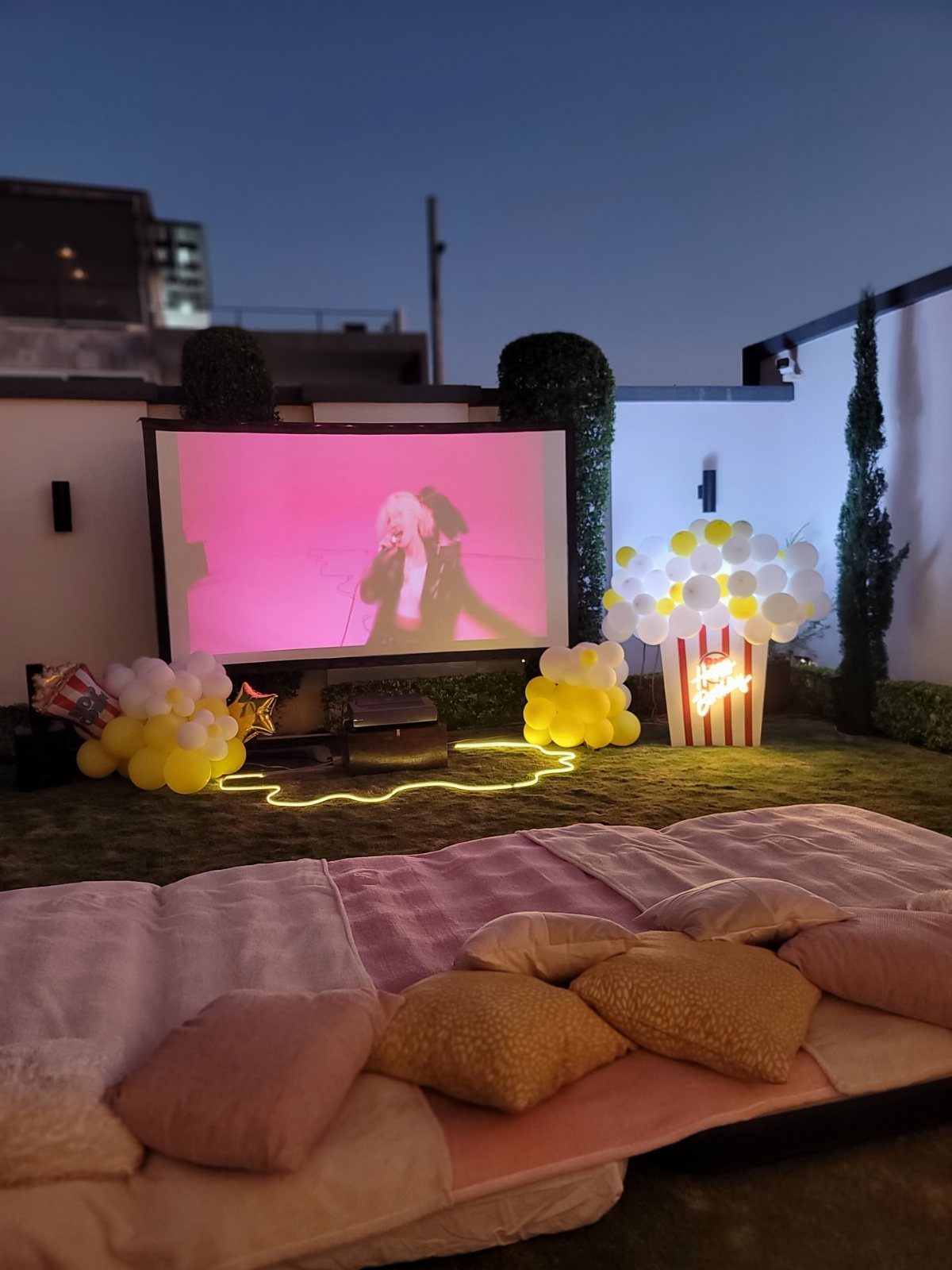 Outdoor movie screen with floor mattress, pillows, and balloons on a green lawn