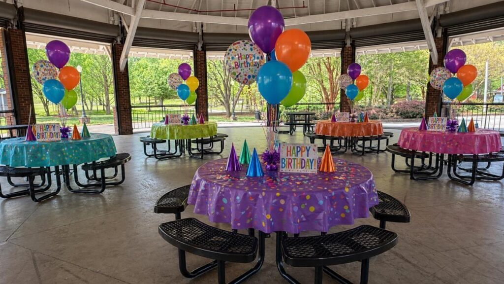Colorful birthday party table setups with balloons in a covered outdoor pavilion.
